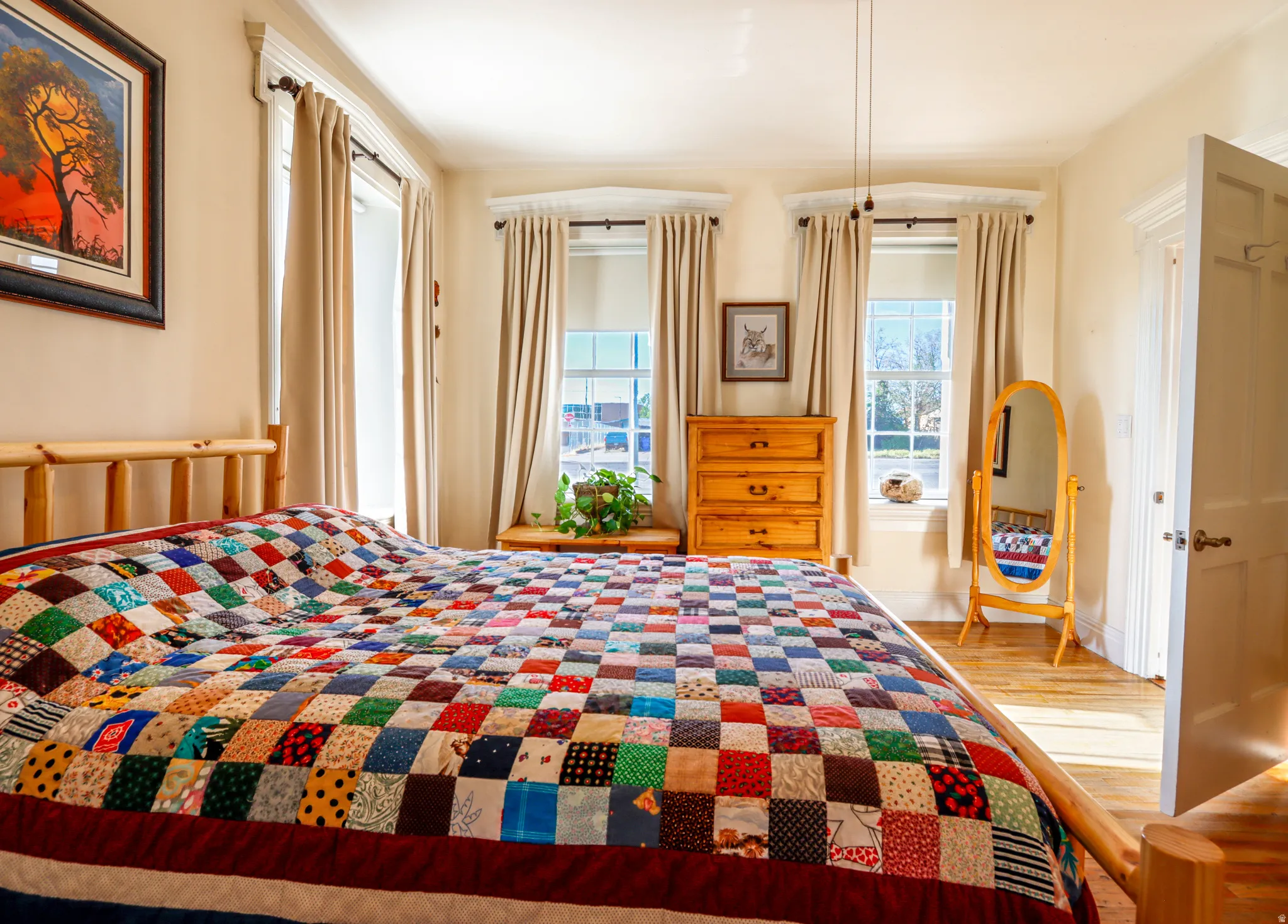 Bedroom with wood-finish flooring, featuring two large windows with full drapes, a wood bed frame, light-colored walls, and a white paneled door