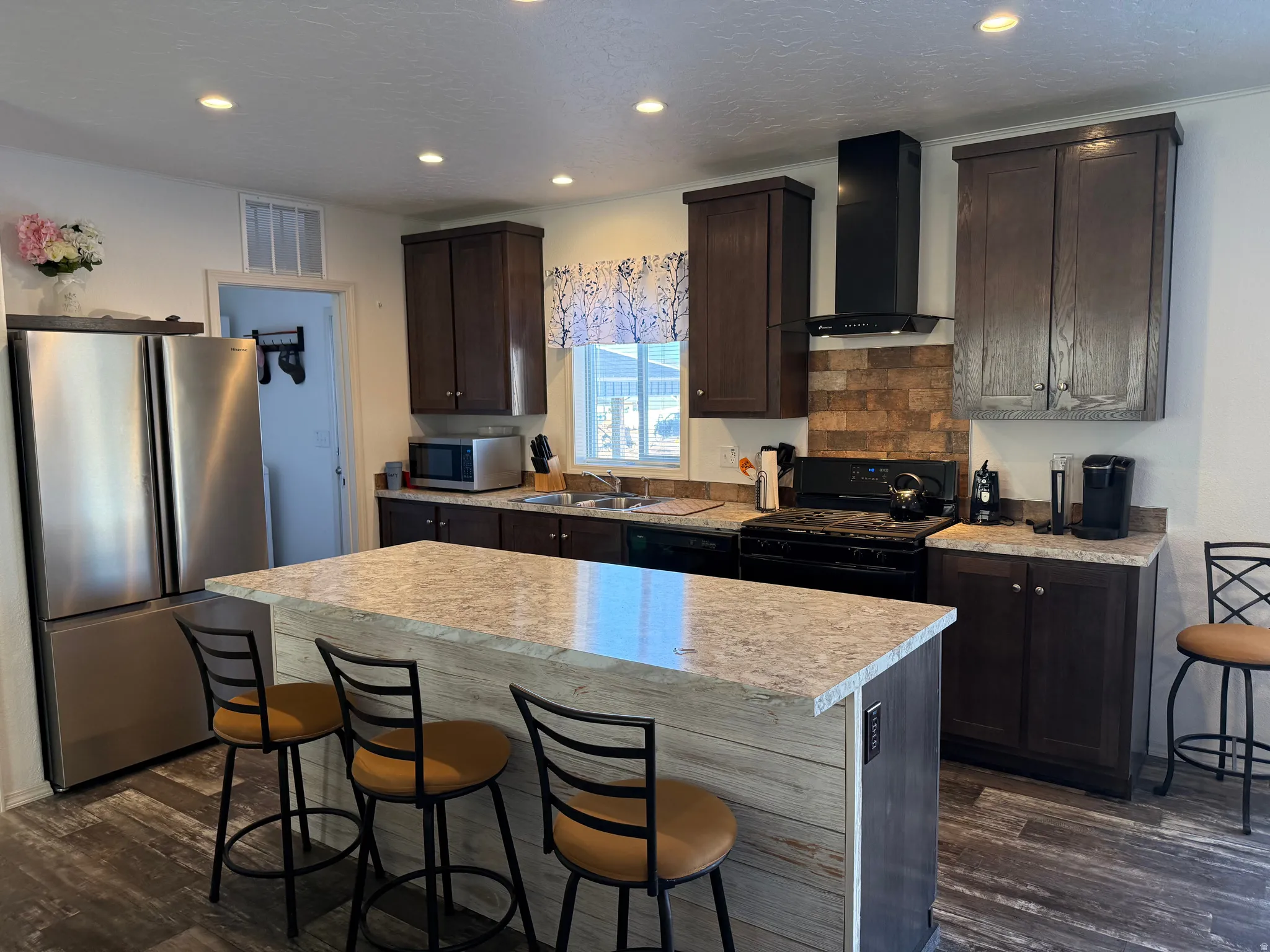 Kitchen featuring a kitchen breakfast bar, black appliances, wall chimney range hood, a kitchen island, and dark brown cabinetry