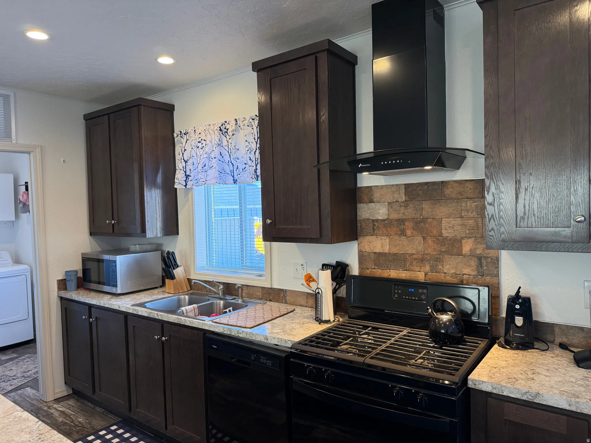 Kitchen featuring dark brown cabinetry, black appliances, light countertops, wall chimney range hood, and recessed lighting
