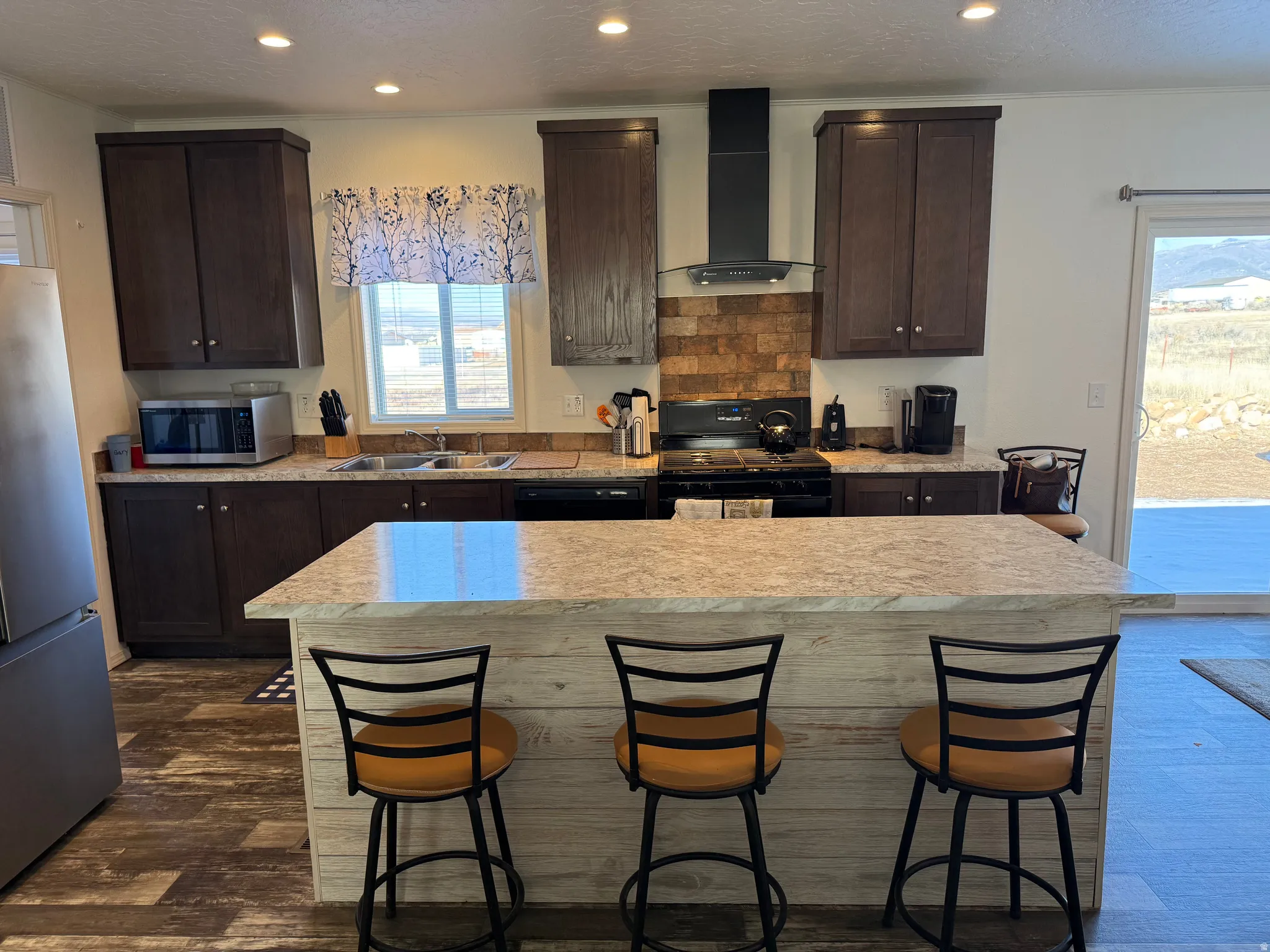 Kitchen featuring a textured ceiling, dark brown cabinets, wall chimney range hood, a center island, and recessed lighting