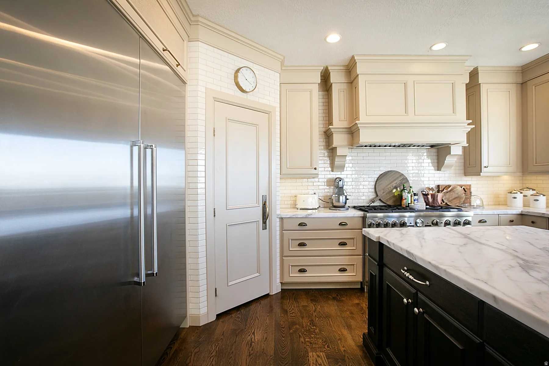 Kitchen with cream cabinets, dark cabinetry on the island, stainless steel built in refrigerator, Calcutta marble counters, and dark wood finished floors