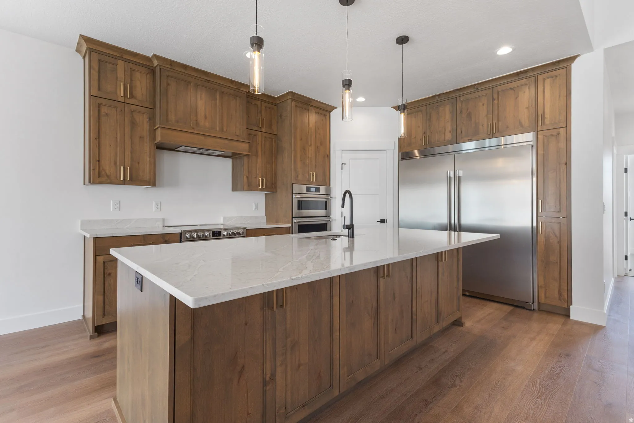 Kitchen featuring stainless steel appliances, pendant lighting, light stone countertops, an island with sink, and light wood finished floors