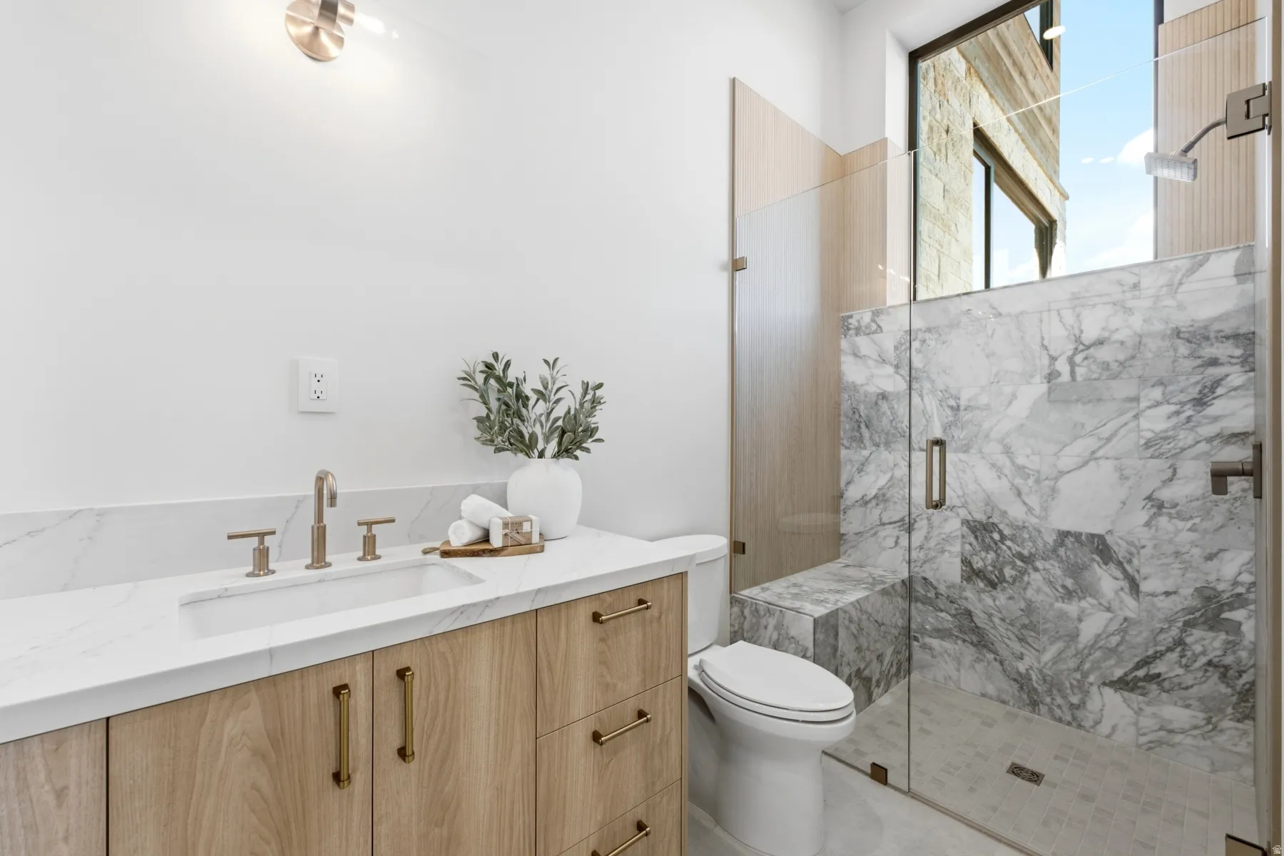 Bathroom featuring a wood-finish vanity with an integrated sink and brushed brass fixtures, a marble-patterned countertop, and a glass-enclosed shower with a built-in bench and a large transom window