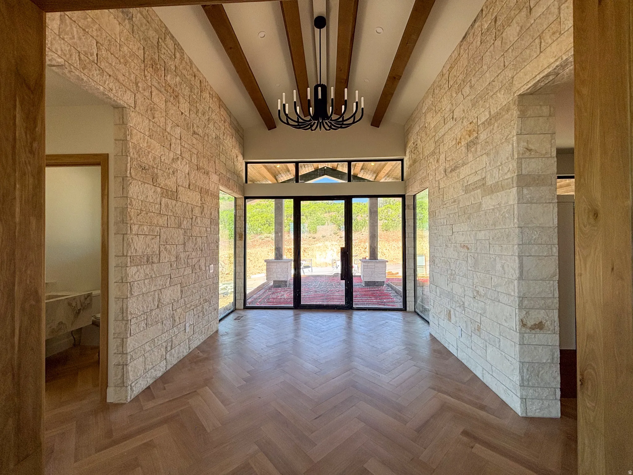 Entryway featuring herringbone wood-finish flooring, stone-clad walls, exposed wood ceiling beams, a black candelabra chandelier, and floor-to-ceiling windows
