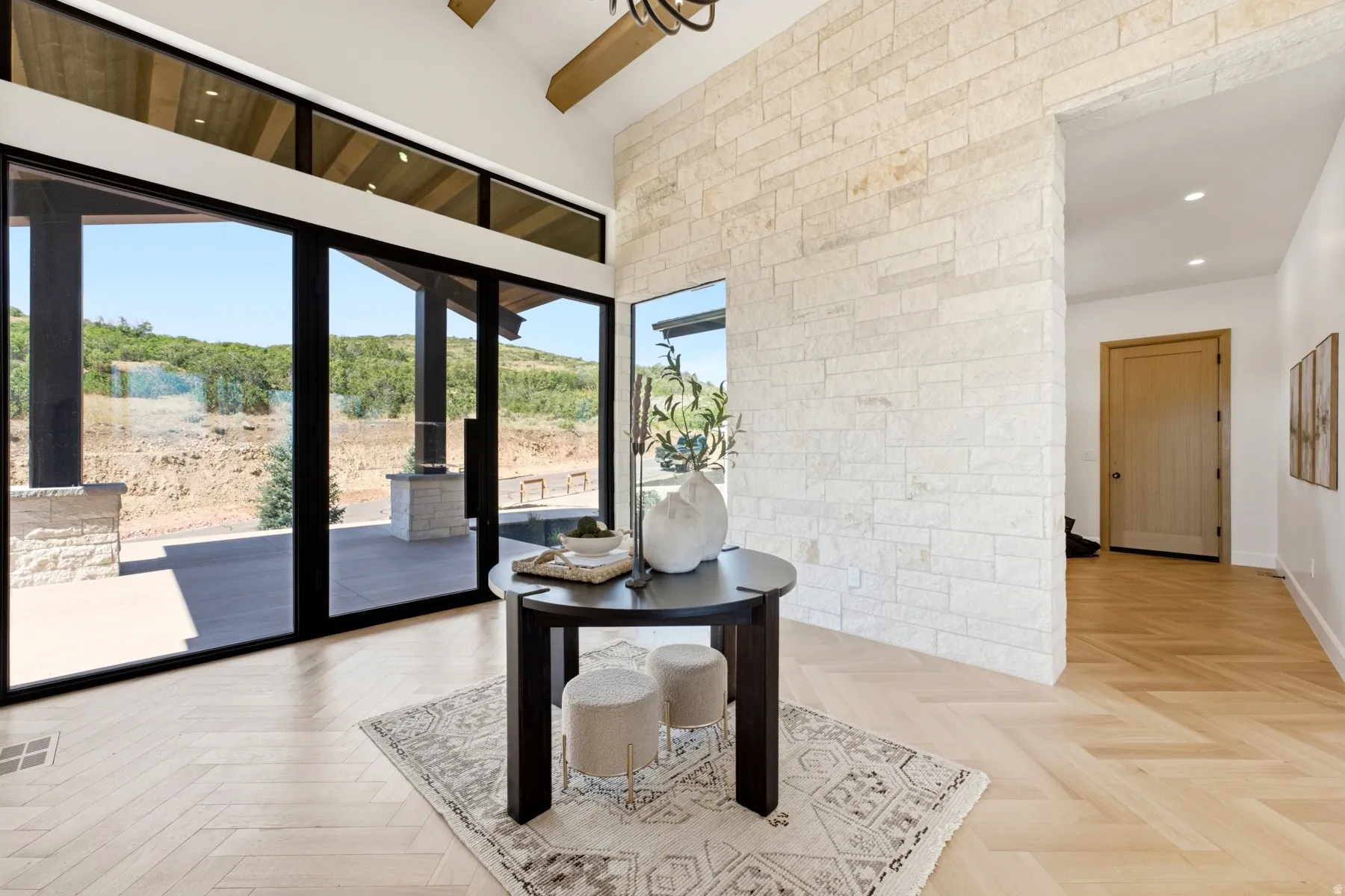 Foyer featuring extensive glass doors, light wood-finish flooring in a herringbone pattern, a stone accent wall, and a high vaulted ceiling with exposed wood beams