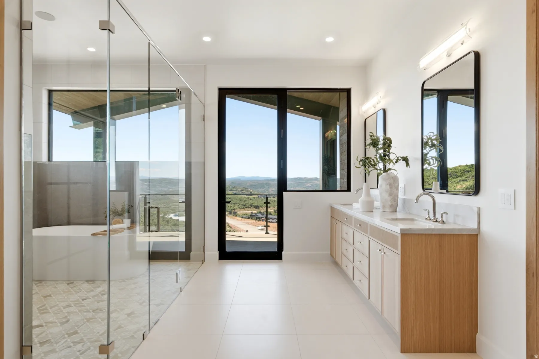 Dual vanity with undermount sinks and light wood-finish cabinetry, floor-to-ceiling glass shower enclosure, freestanding soaking tub, and large format floor tiling