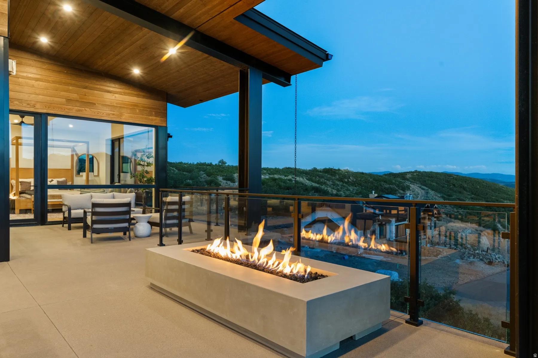 Spacious outdoor patio with a built-in fire pit, glass railing, wood-paneled ceiling, and recessed lighting