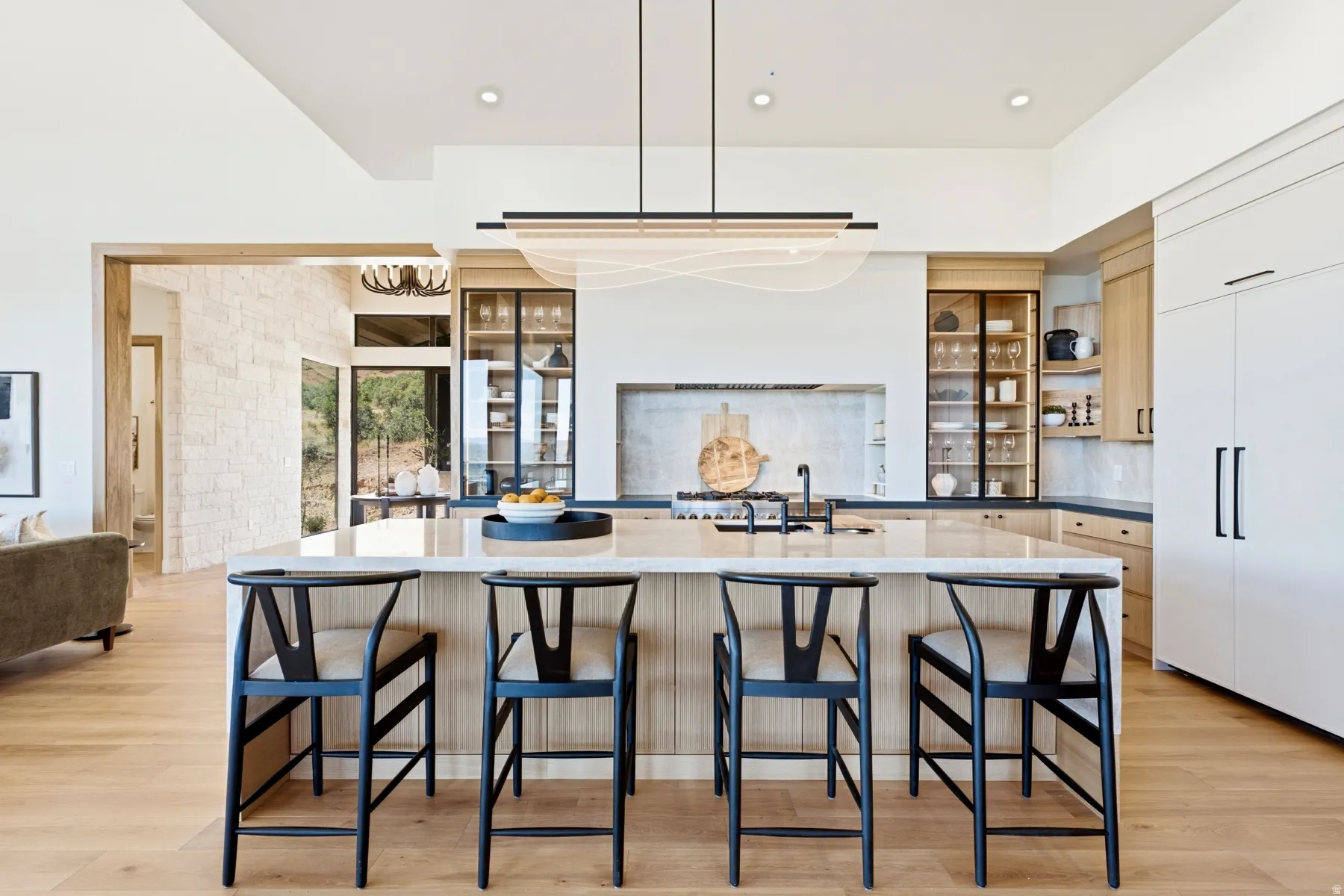 Spacious kitchen featuring a large central island with seating, integrated appliances, natural wood-finish cabinetry with glass-front display sections, a full-height tile backsplash, and a contemporary linear pendant light fixture