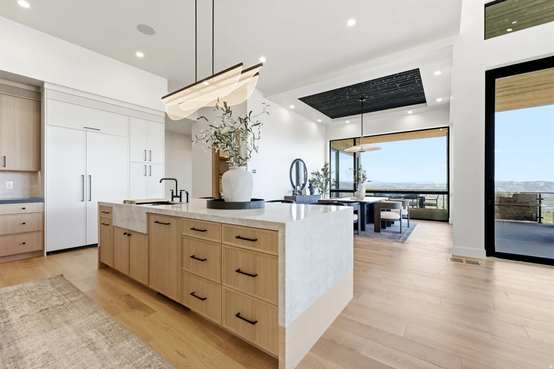 Open-concept kitchen and dining area featuring a spacious island with a farmhouse sink, light wood-finish flooring, full-height white cabinetry, and extensive window walls offering distant landscape scenery