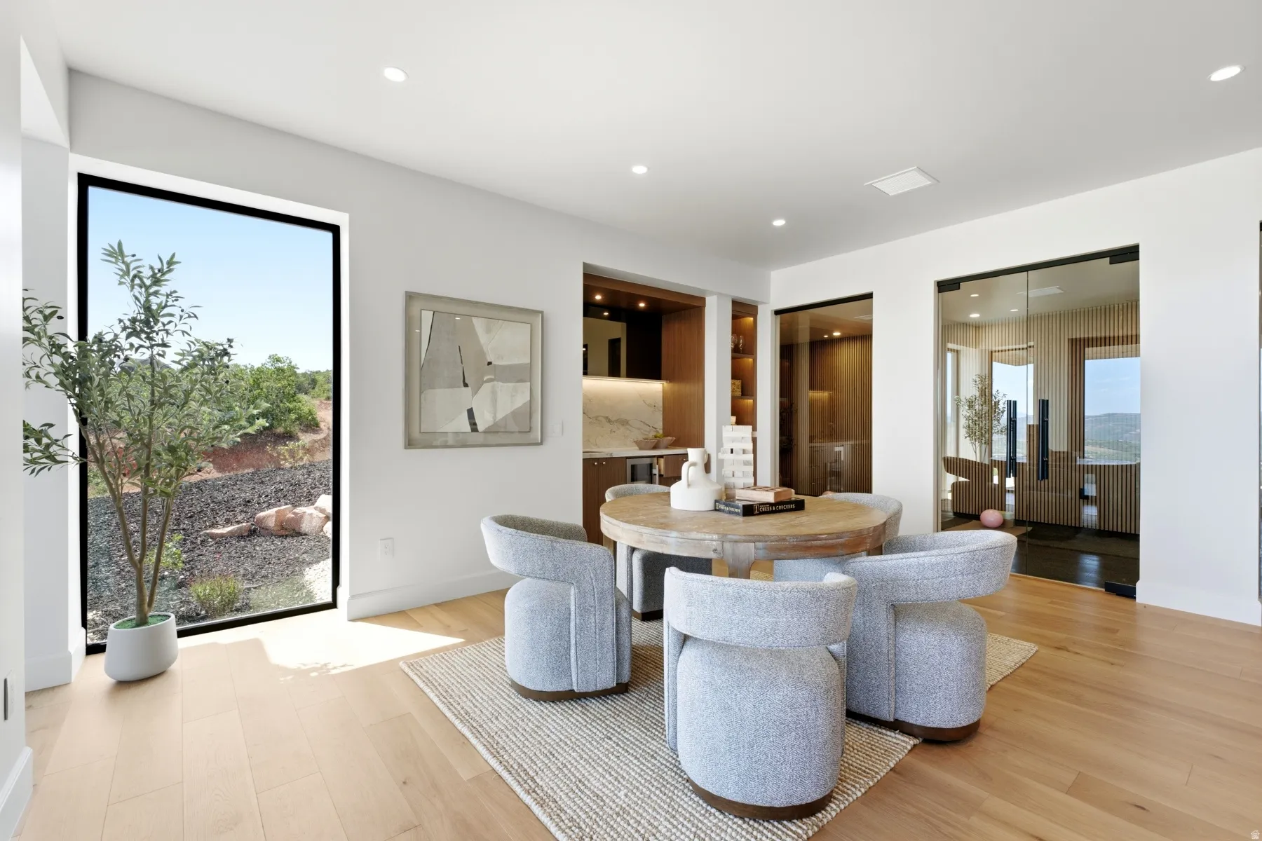 Spacious interior featuring light wood-finish flooring, a large floor-to-ceiling window, a built-in wet bar with shelving, and two glass-paneled entryways with black frames