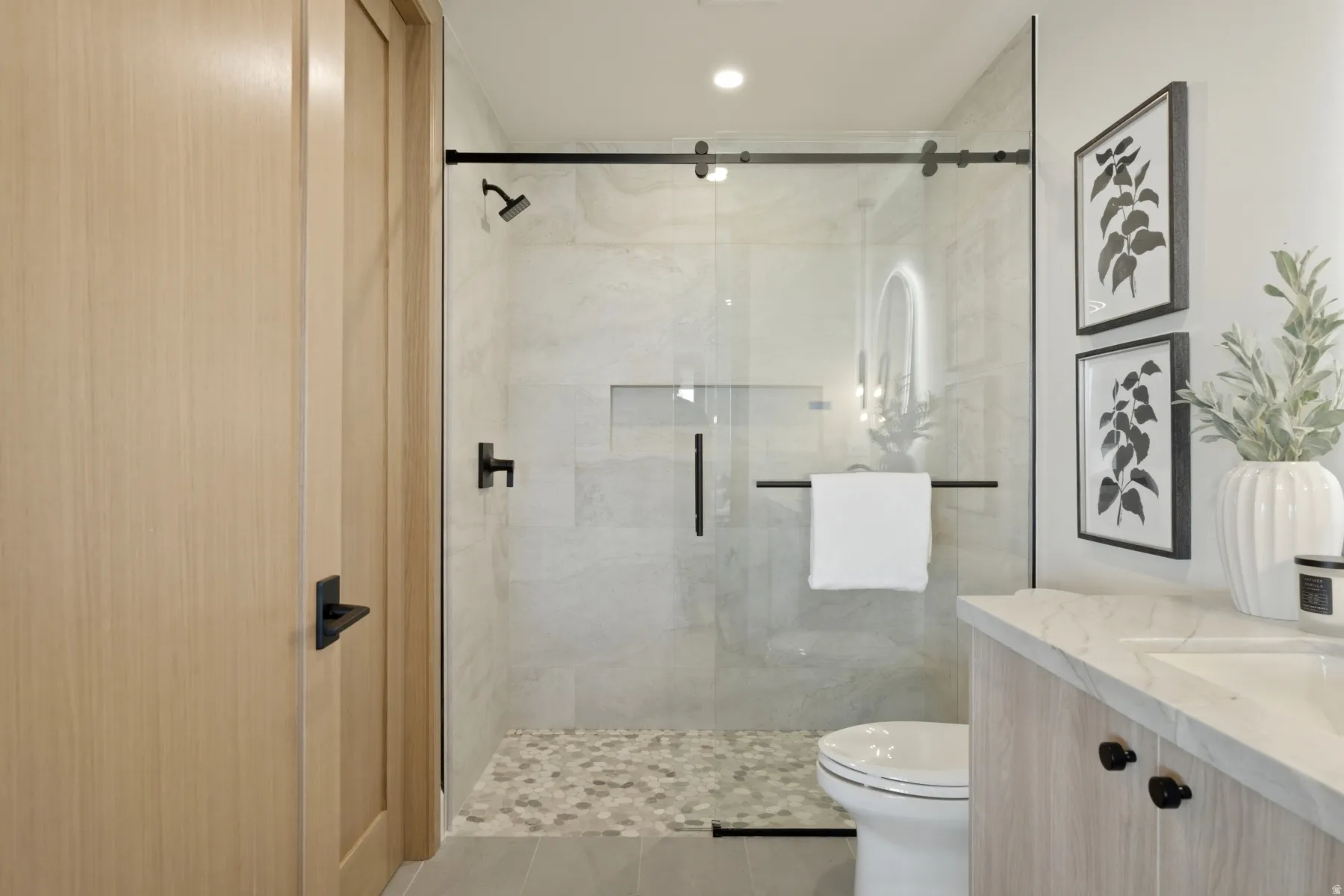 Frameless glass shower enclosure with matte black hardware, pebble-tiled shower floor, integrated shower niche, and a light wood-finish vanity with a stone countertop