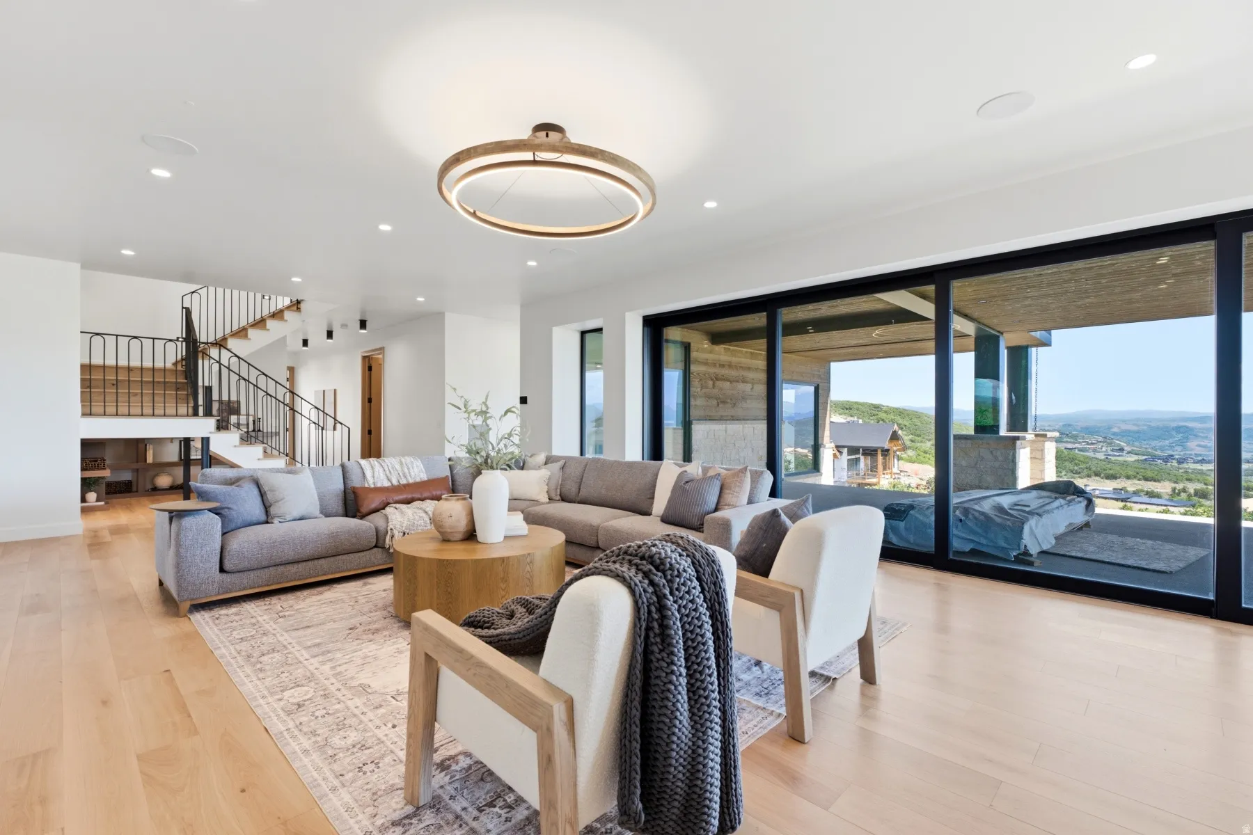 Spacious living area featuring light wood-finish flooring, a contemporary chandelier, recessed lighting, and a prominent staircase with black metal railings