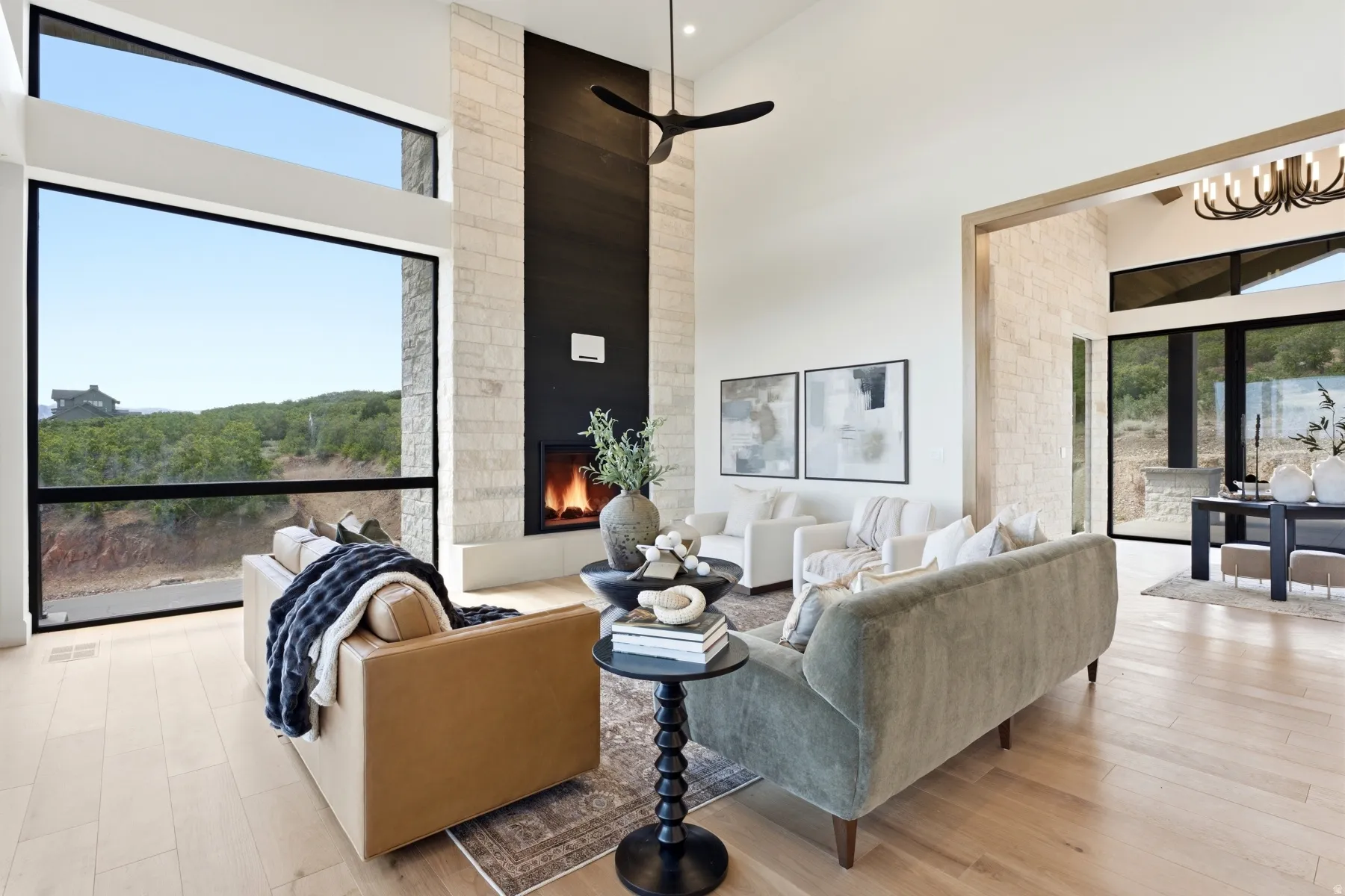 Spacious open-concept living area featuring a floor-to-ceiling stone fireplace, expansive windows, wood-finish flooring, and high ceilings