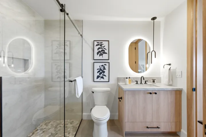 Glass-enclosed shower with a tiled interior, single vanity with a light wood finish, oval-shaped backlit mirror, and matte black fixtures