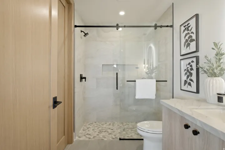 Frameless glass shower enclosure with matte black hardware, pebble-tiled shower floor, integrated shower niche, and a light wood-finish vanity with a stone countertop