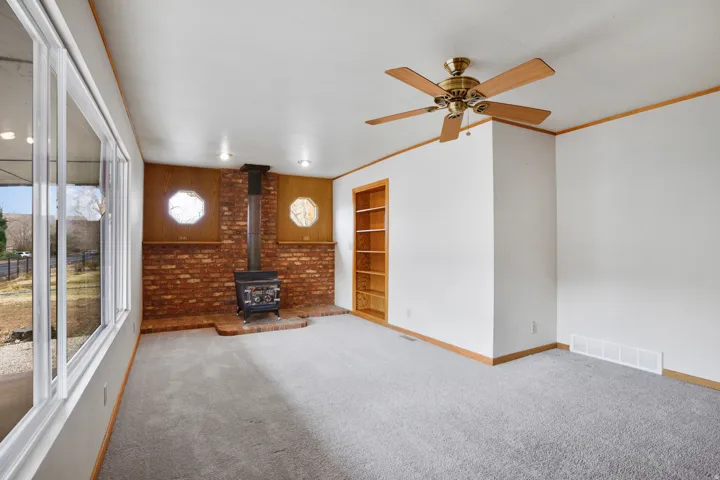 Unfurnished living room with a wood stove, carpet, built in shelves, a ceiling fan, and ornamental molding