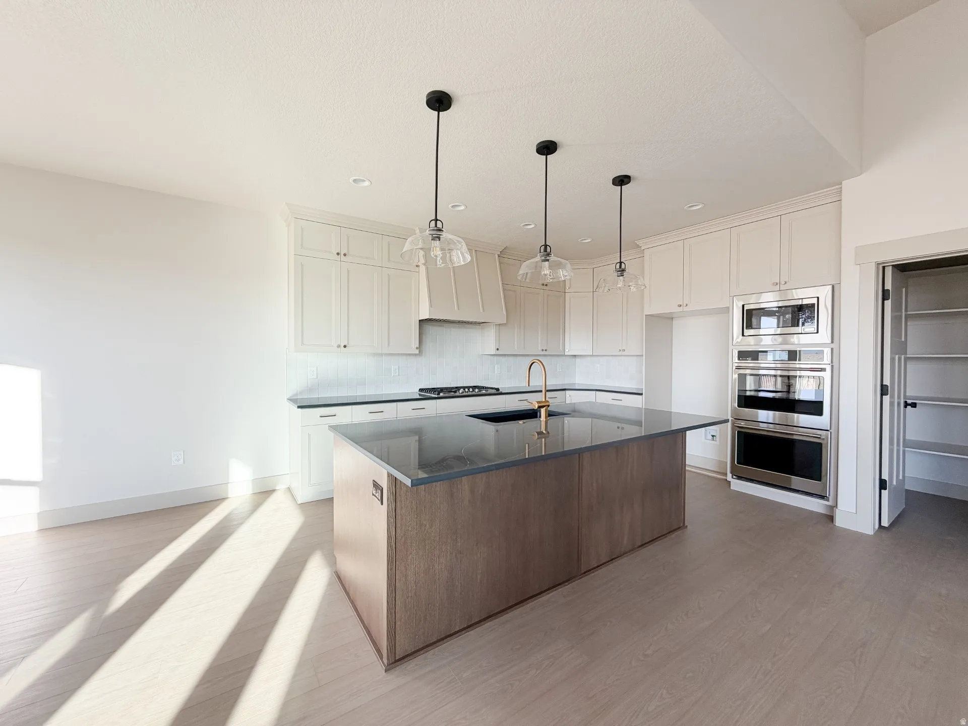 Kitchen featuring light wood-style floors, appliances with stainless steel finishes, a kitchen island with sink, decorative light fixtures, and white cabinets