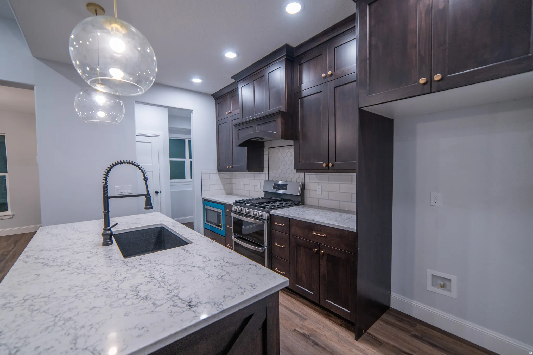 Kitchen featuring appliances with stainless steel finishes, pendant lighting, dark wood finished floors, light stone counters, and dark brown cabinetry