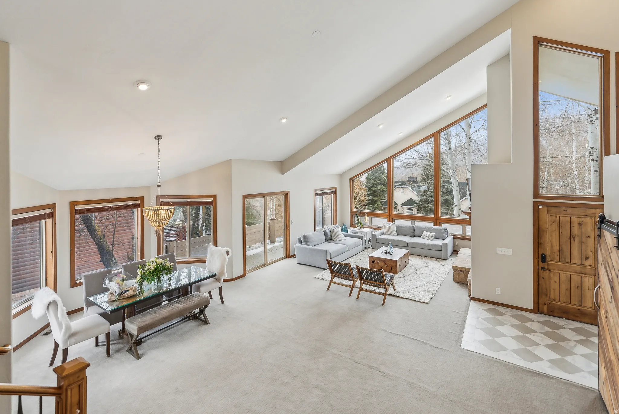 Soaring vaulted ceilings with recessed lighting, expansive floor-to-ceiling windows, a sliding glass door for outdoor access, a distinct dining area with a chandelier, and a wood-finish barn door
