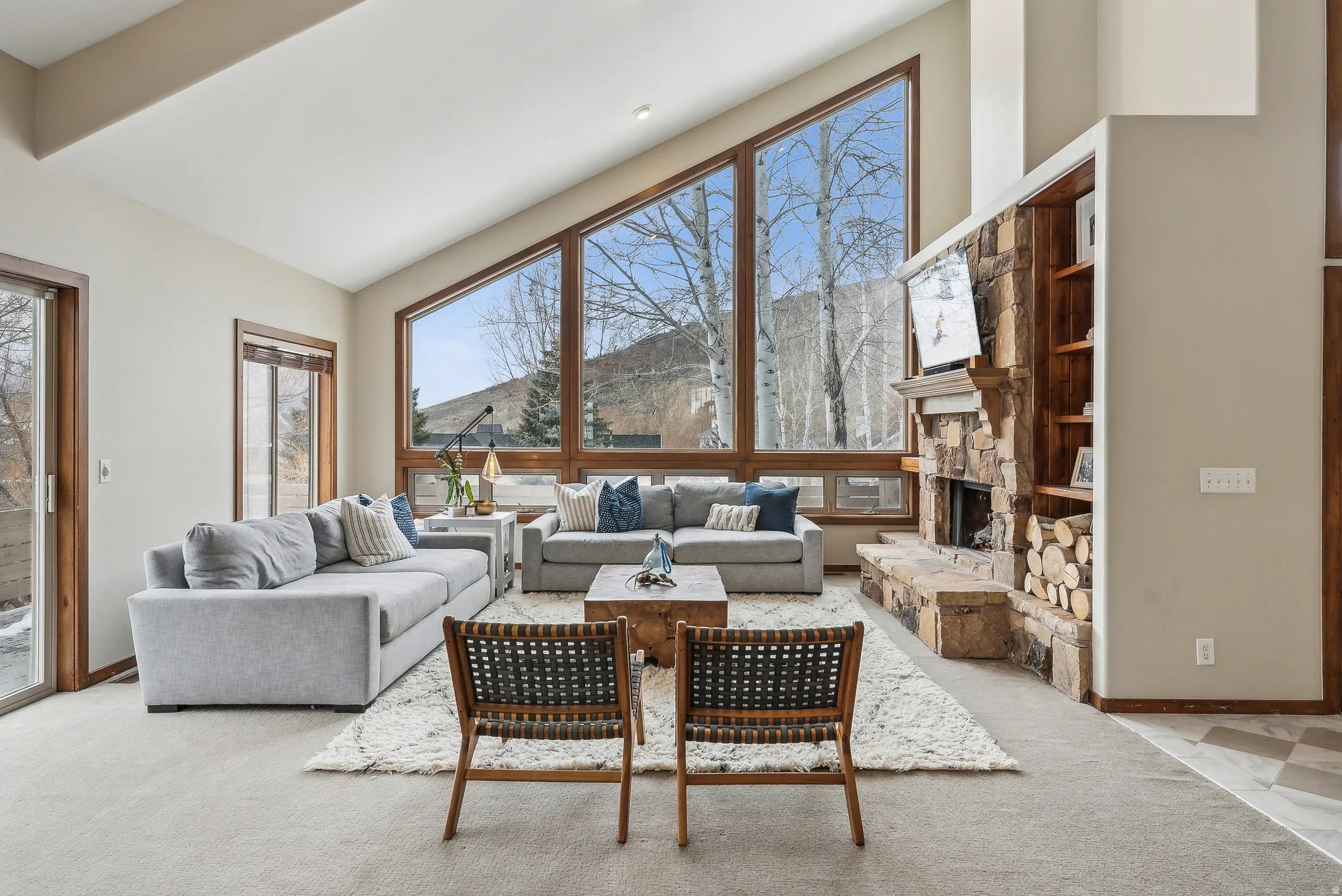 Vaulted ceiling with extensive window fenestration, stone-clad fireplace with a hearth, built-in wood shelving, and wood-finish sliding glass doors