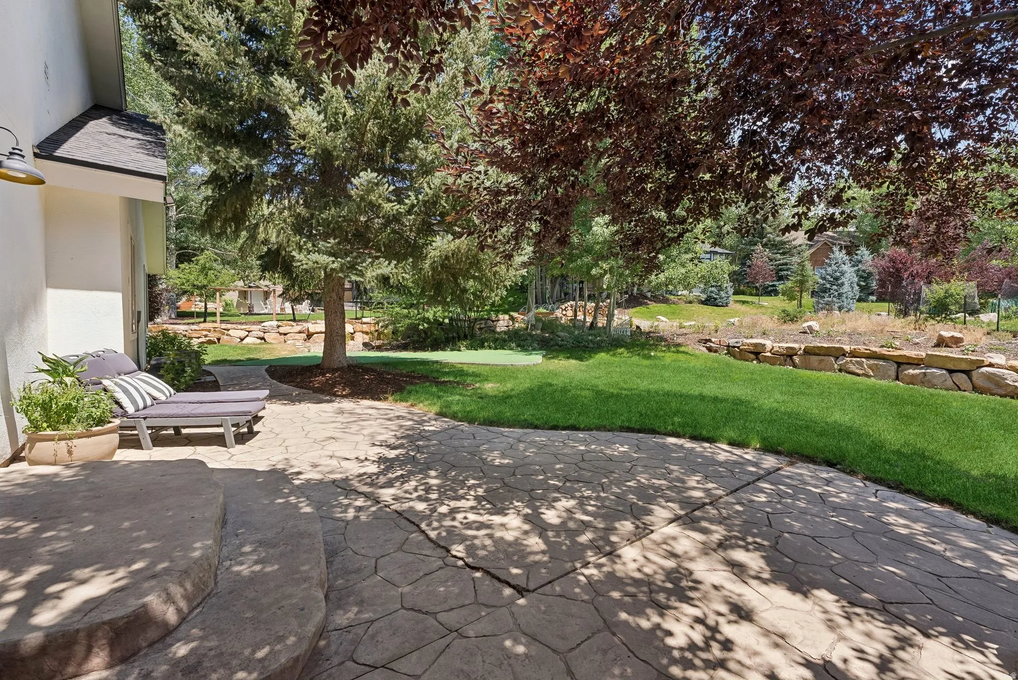 Textured patio surface with multiple steps, expansive lawn, mature shade trees, and a stone retaining wall