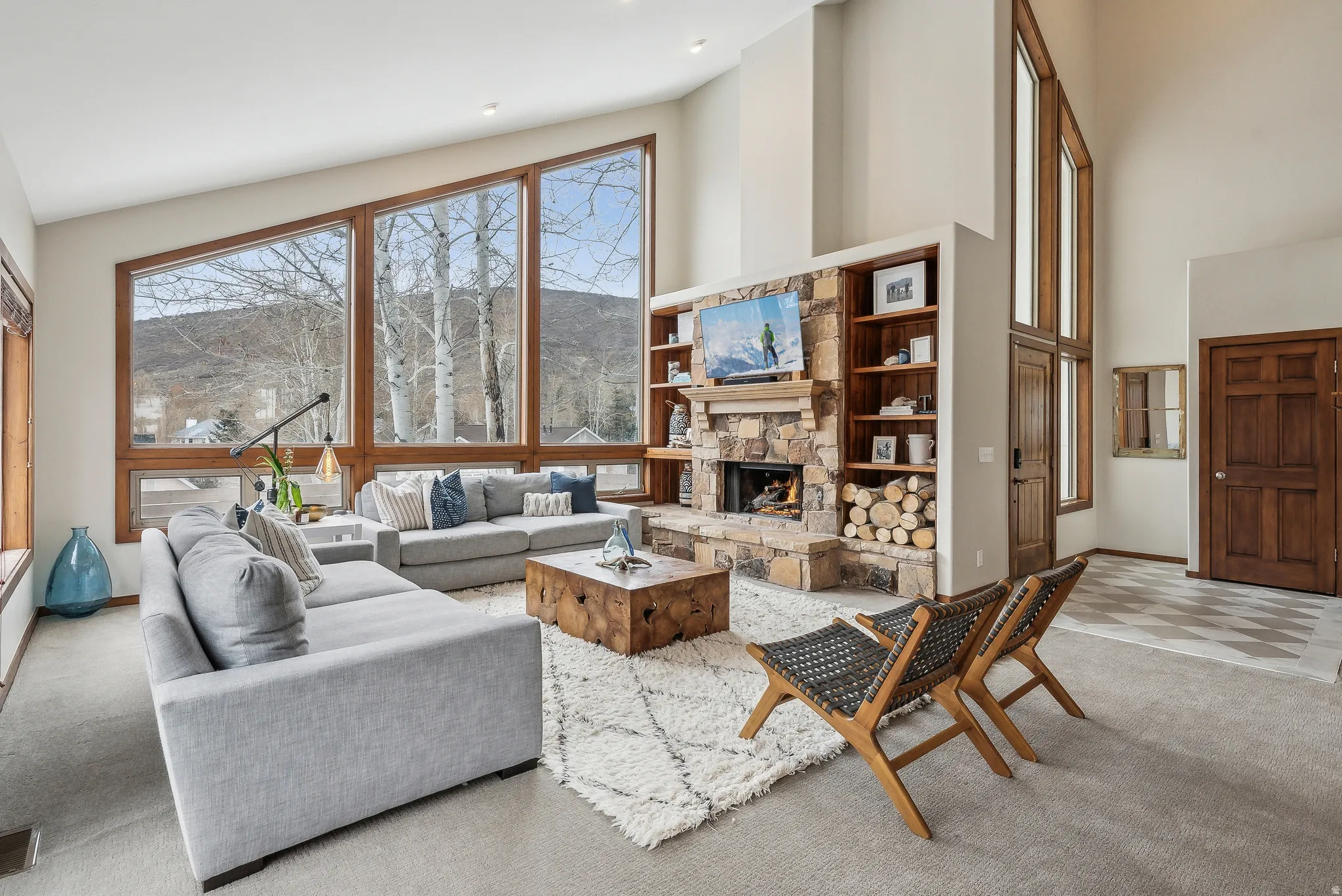 Vaulted ceilings with expansive window walls, stone-clad fireplace with wood storage, built-in shelving, and wood-paneled entry door