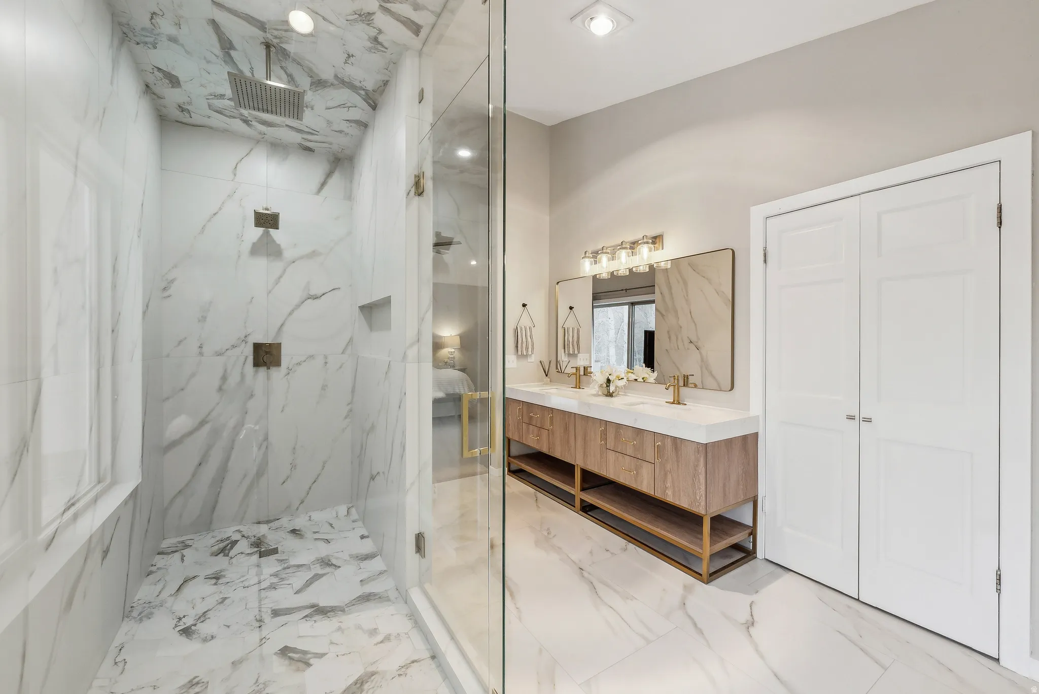 Spa-inspired primary bathroom featuring marble-clad shower enclosure with frameless glass partition, marble flooring throughout, floating wood-finish vanity with dual sinks, integrated mirror with integrated lighting, recessed ceiling lights, and polished