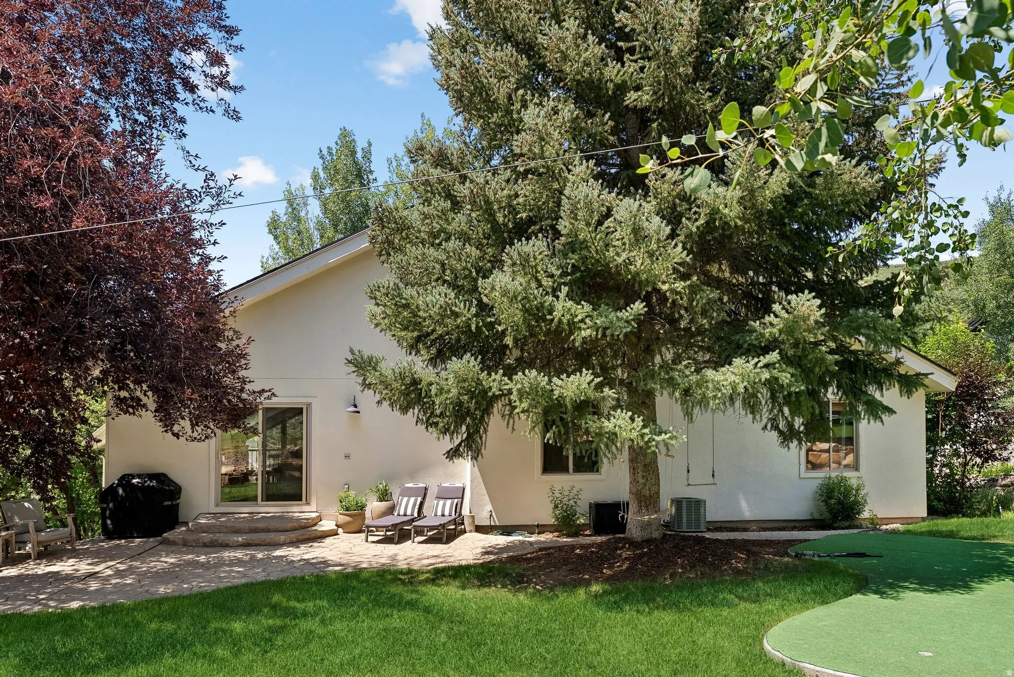 Rear exterior with a stone patio, sliding glass door access, an expansive lawn, mature foliage, and a putting green