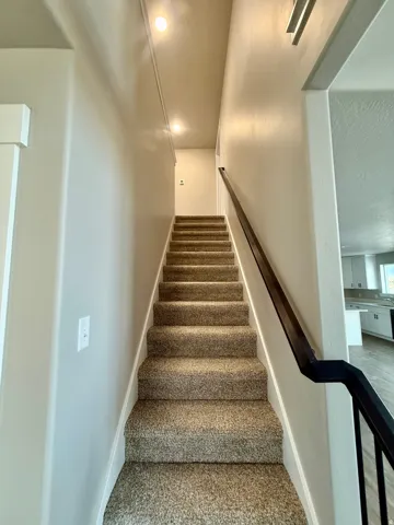 Carpeted staircase featuring a dark-finish handrail and recessed lighting