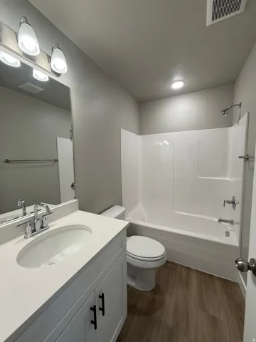 White vanity with a light-toned countertop, wood-finish flooring, full-size mirror, and a white tub with shower