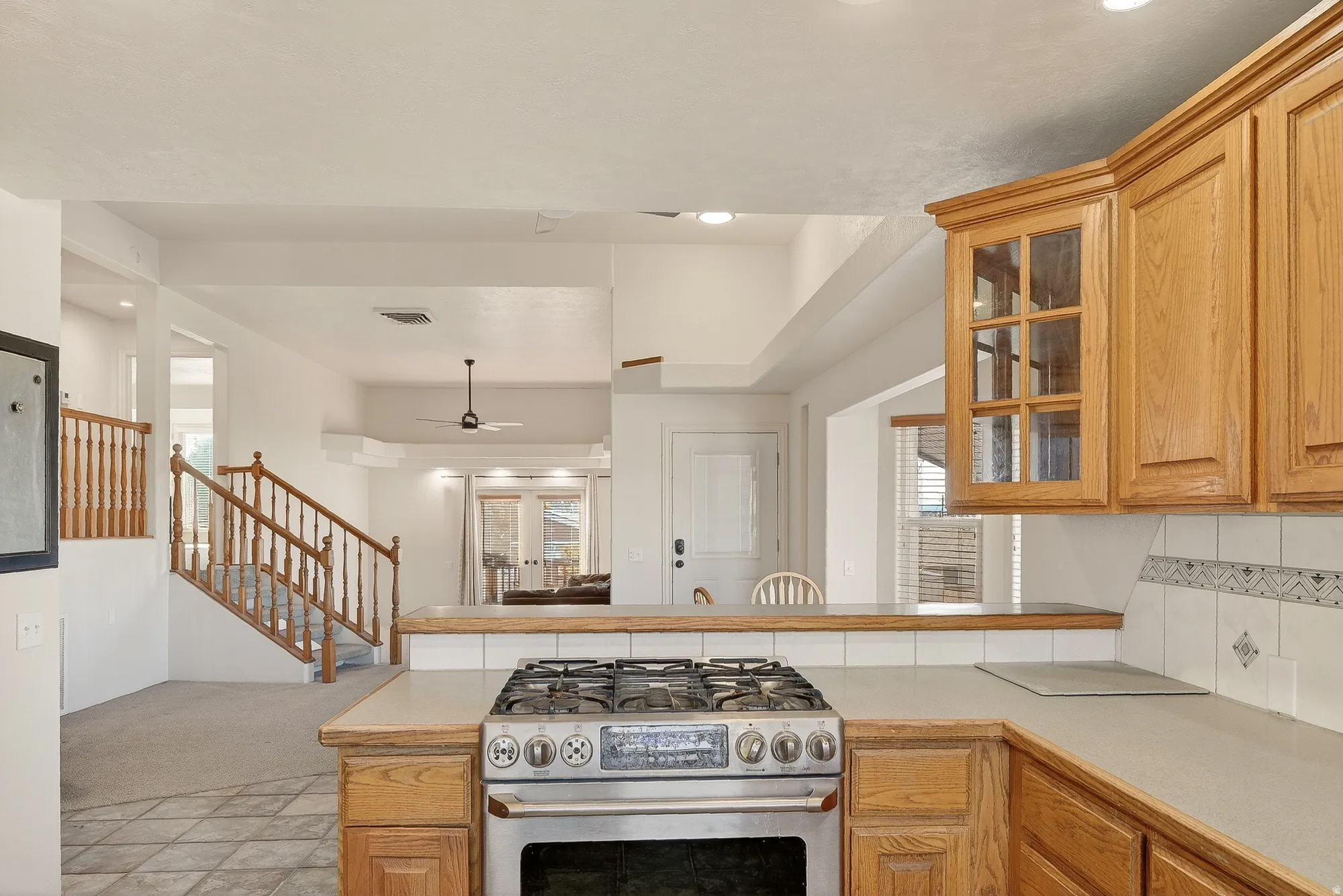 Kitchen with light countertops, gas stove, brown cabinetry, glass insert cabinets, and recessed lighting