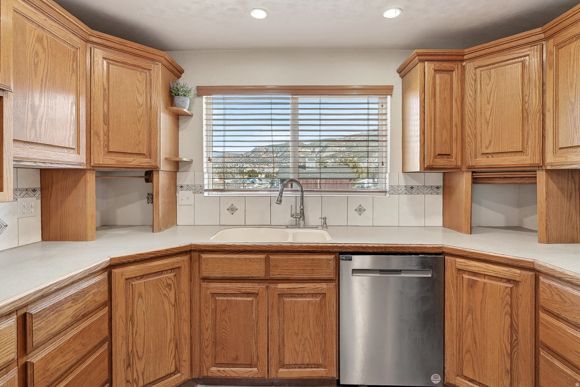 Kitchen featuring light countertops, stainless steel dishwasher, open shelves, brown cabinetry, and recessed lighting