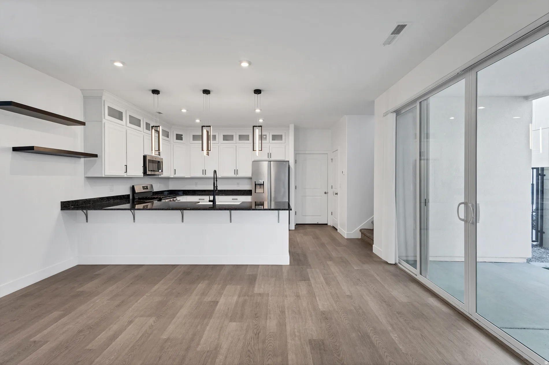 Open-concept living area featuring wood-finish flooring, a kitchen with white cabinetry and dark countertops, stainless steel appliances, and a glass sliding door leading to an outdoor patio