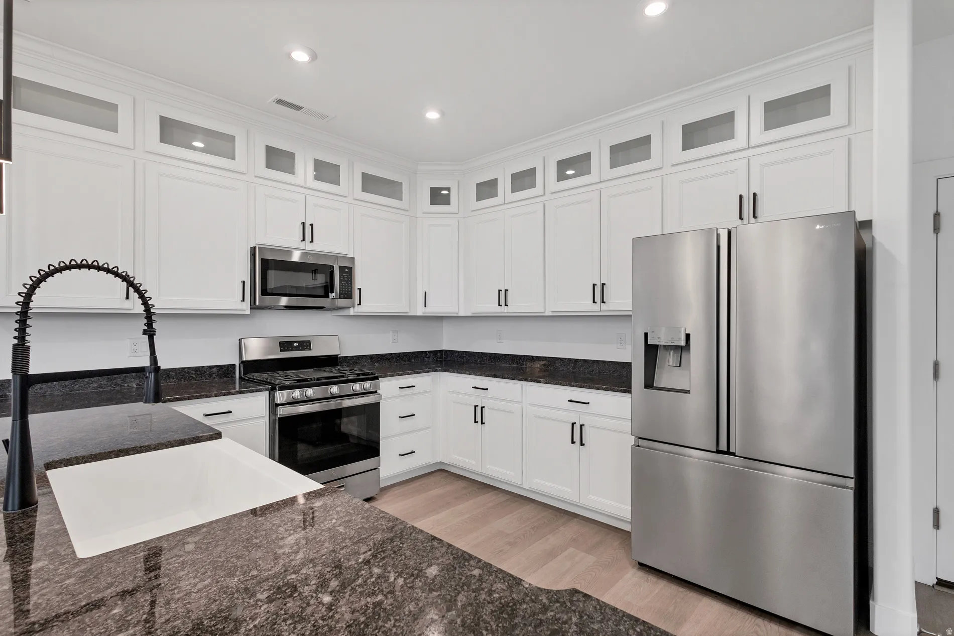 White cabinetry with glass-front uppers, stainless steel appliances, dark stone countertops, wood-finish flooring, and a gooseneck faucet