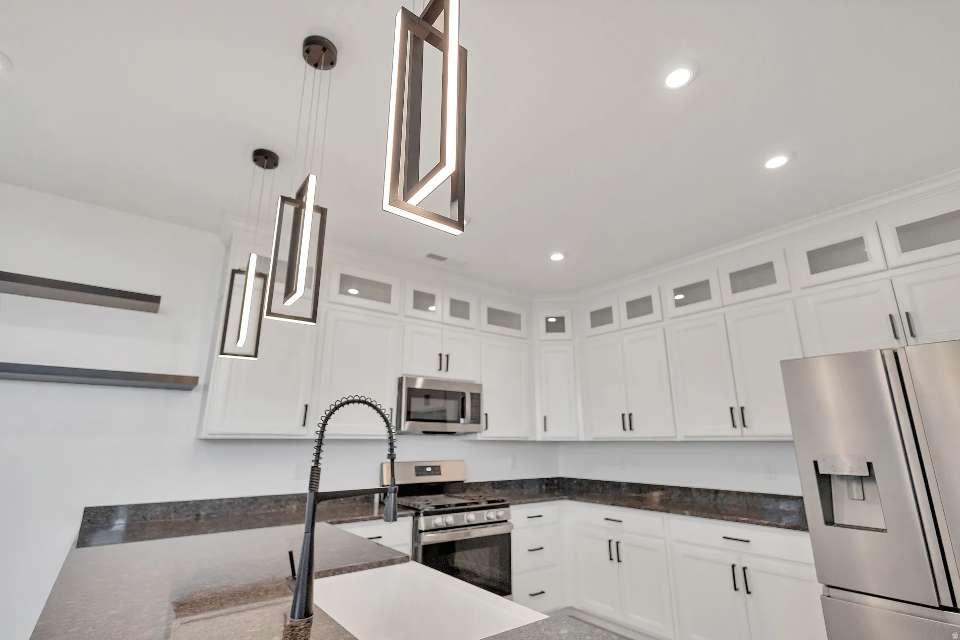 White cabinetry with upper glass-front accents, stainless steel appliances, dark countertops, recessed lighting, and modern pendant light fixtures