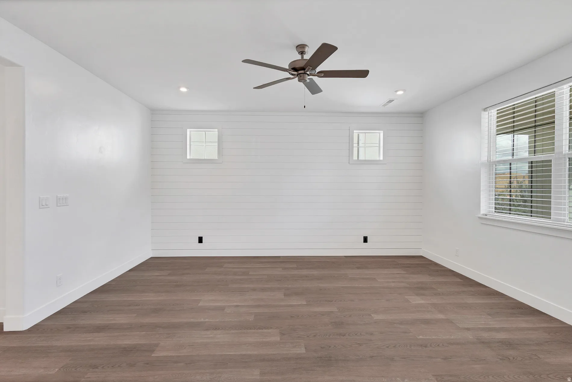 Spacious room featuring wood-finish flooring, white shiplap accent wall, recessed lighting, ceiling fan, and multiple windows
