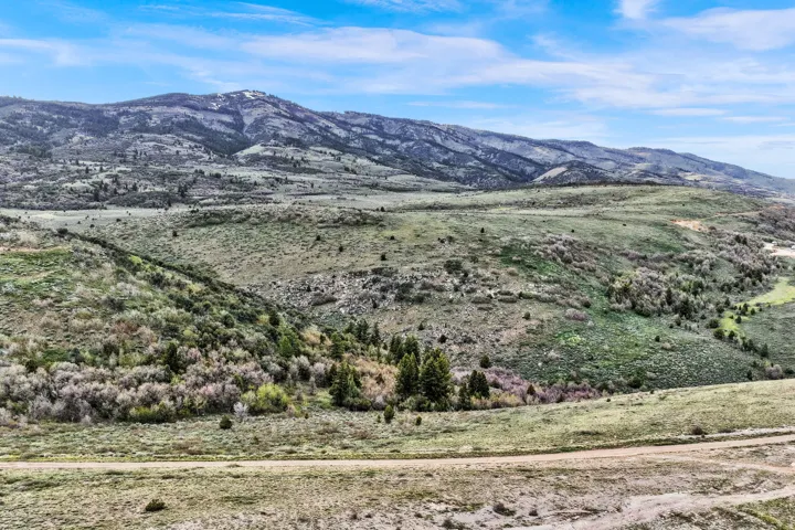 Expansive mountain landscape featuring varied terrain, natural vegetation, distant peaks, a dirt track, and a clear blue sky
