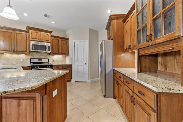 Kitchen with brown cabinetry, light stone counters, glass insert cabinets, appliances with stainless steel finishes, and pendant lighting