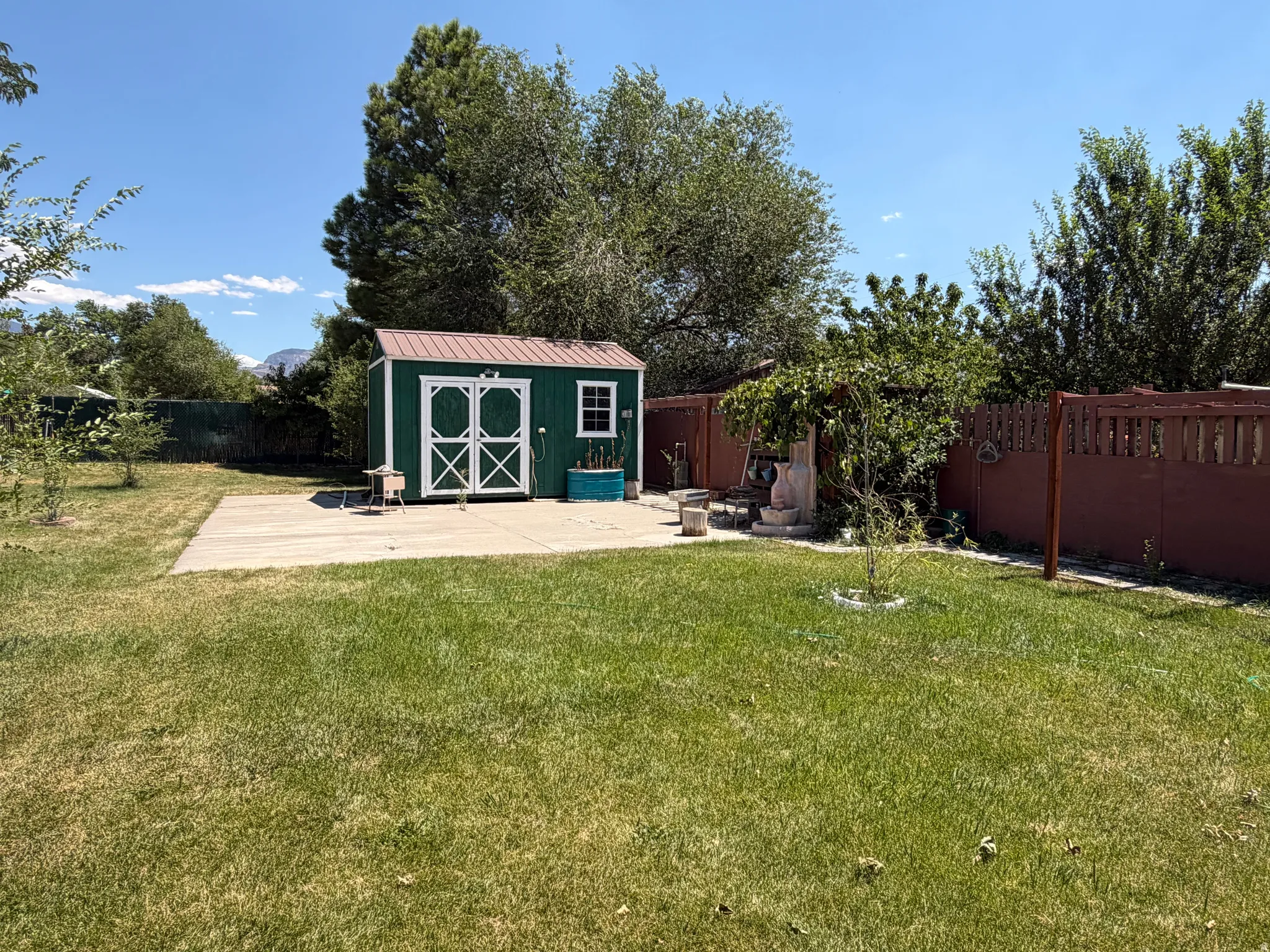 Expansive backyard featuring a green storage shed with a brown metal roof, a concrete patio, and mature perimeter trees