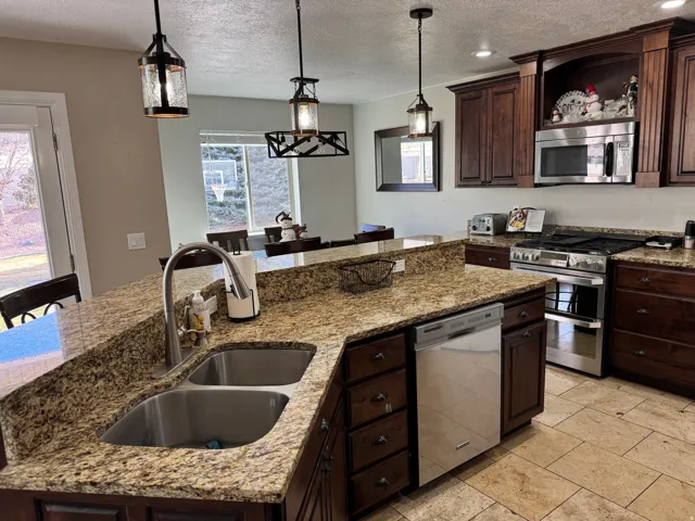 Kitchen featuring dark brown cabinets, appliances with stainless steel finishes, pendant lighting, a textured ceiling, and light stone counters