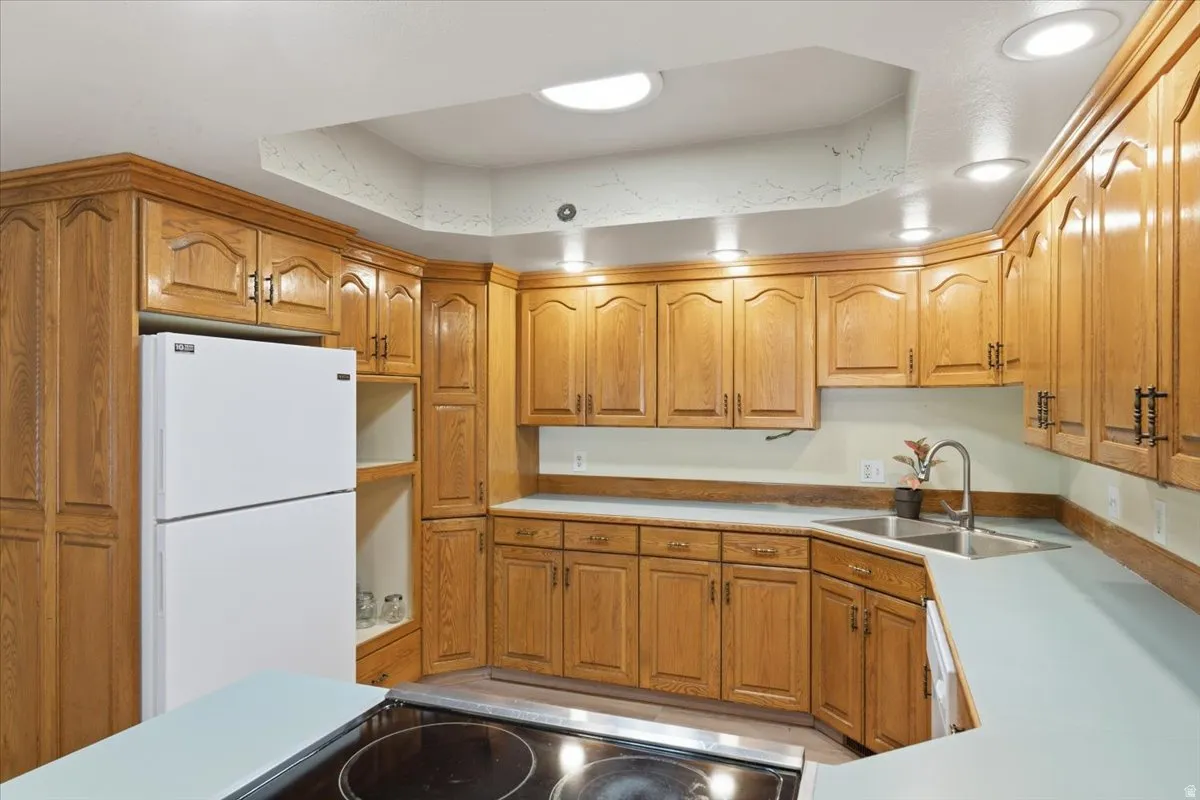 Kitchen with white appliances, a raised ceiling, brown cabinetry, light countertops, and recessed lighting