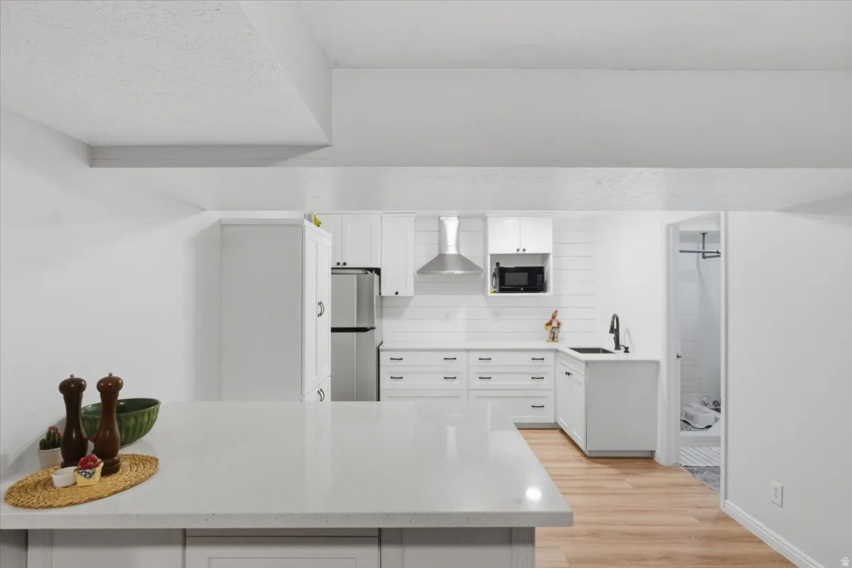 Kitchen with wall chimney exhaust hood, white cabinetry, light wood-type flooring, freestanding refrigerator, and light stone counters
