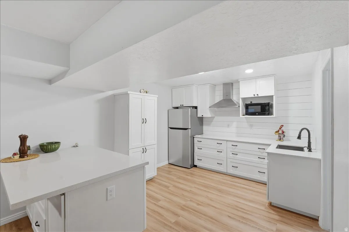 Kitchen with light wood finished floors, freestanding refrigerator, wall chimney exhaust hood, white cabinetry, and black microwave