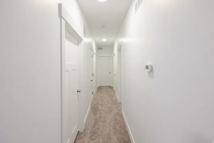 Crisp white walls define this hallway, featuring a textured carpet runner, recessed lighting, white panel doors, and a digital thermostat