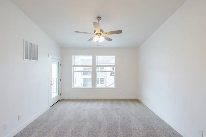 Spacious room featuring carpeted flooring, large windows with blinds, a ceiling fan with integrated lighting, and a glass-paneled door leading to an exterior balcony