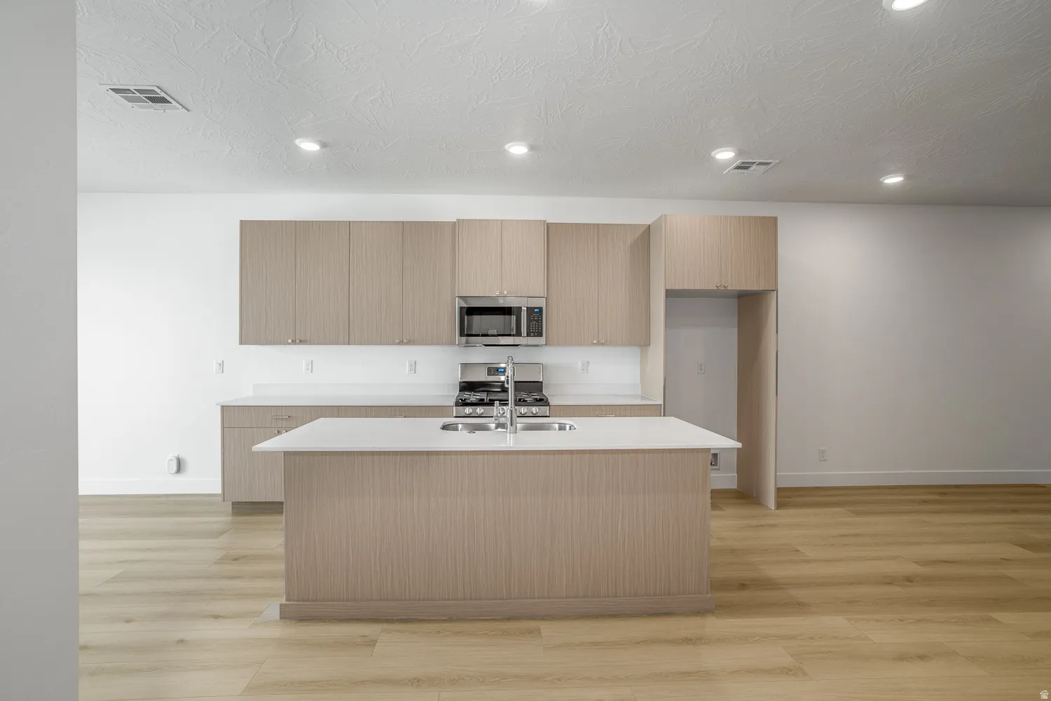 Kitchen with light wood-type flooring, modern cabinets, appliances with stainless steel finishes, light stone counters, and a center island with sink