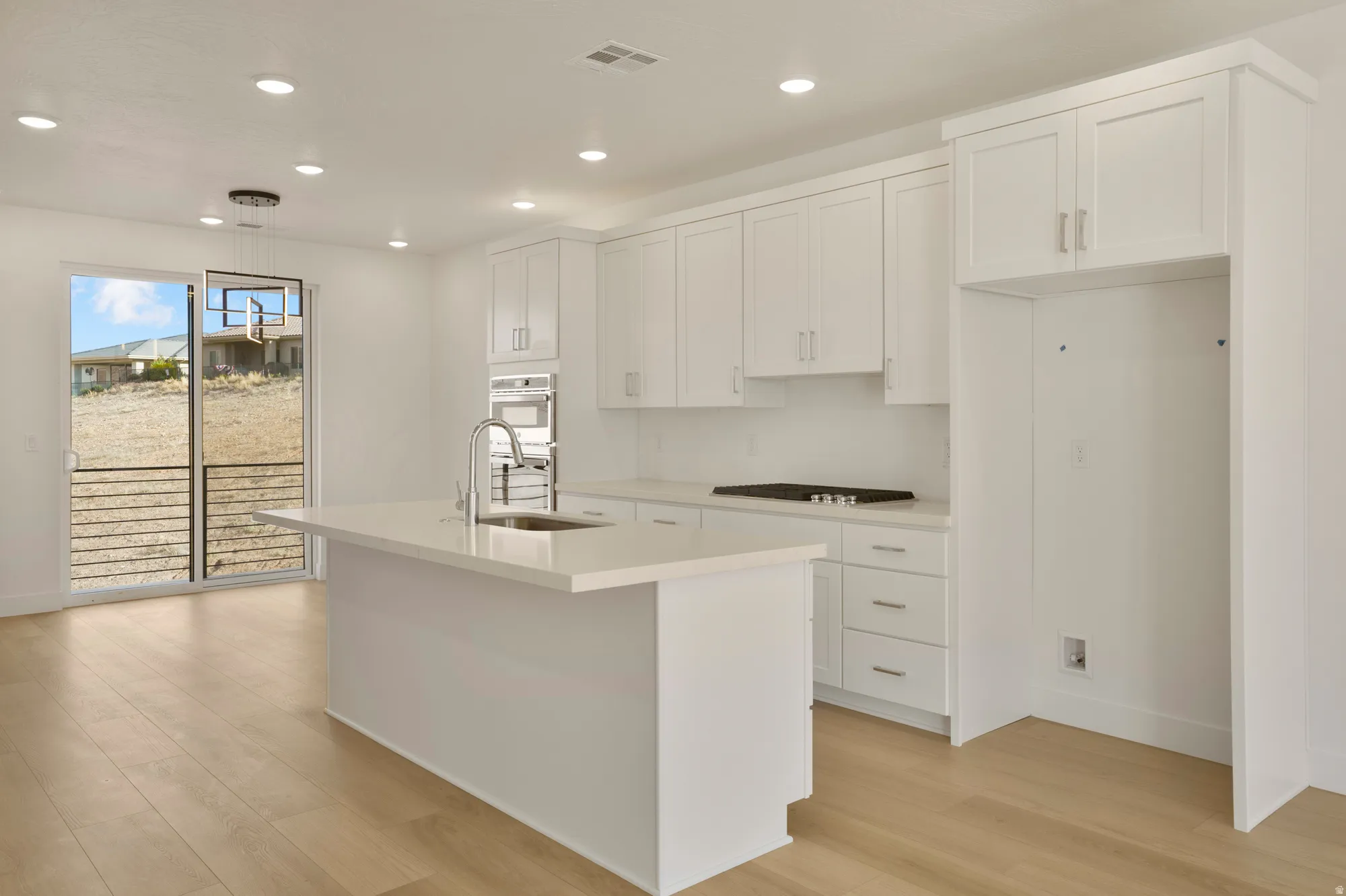 Bright kitchen featuring white shaker cabinetry, an island with an integrated sink, a built-in oven, a cooktop, and wood-finish flooring