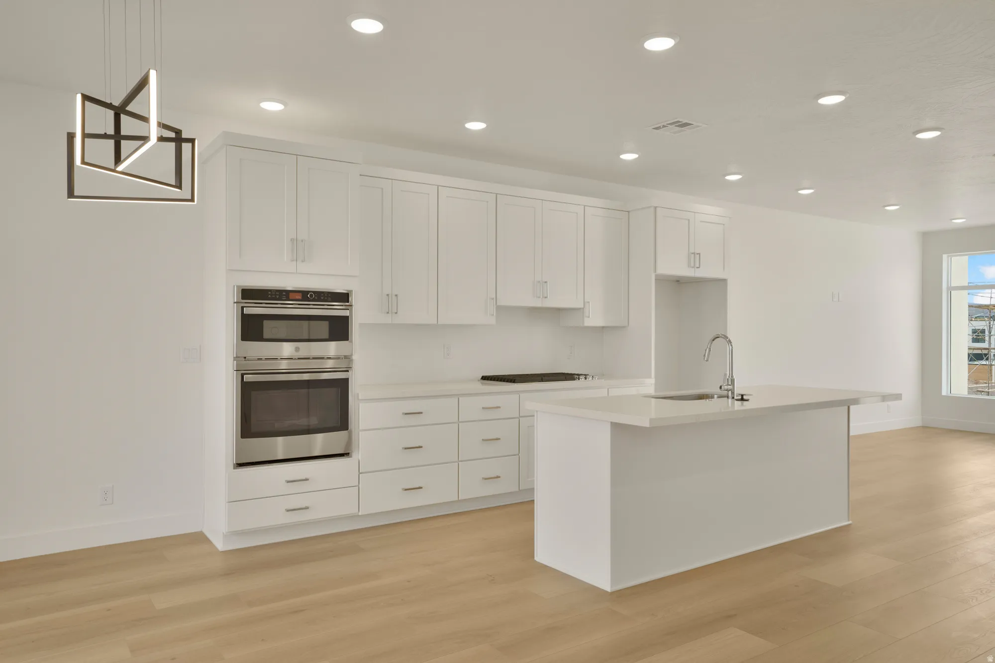 Modern kitchen featuring white shaker-style cabinetry, a built-in stainless steel oven and microwave, light-toned wood-finish flooring, a contemporary island with an integrated sink, and recessed ceiling lighting