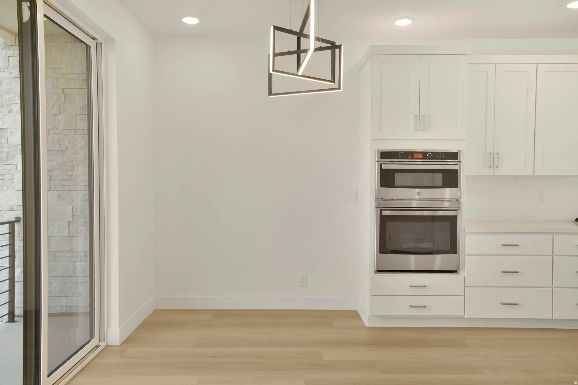 Contemporary kitchen area featuring stainless steel double ovens, white cabinetry, and light wood-finish flooring