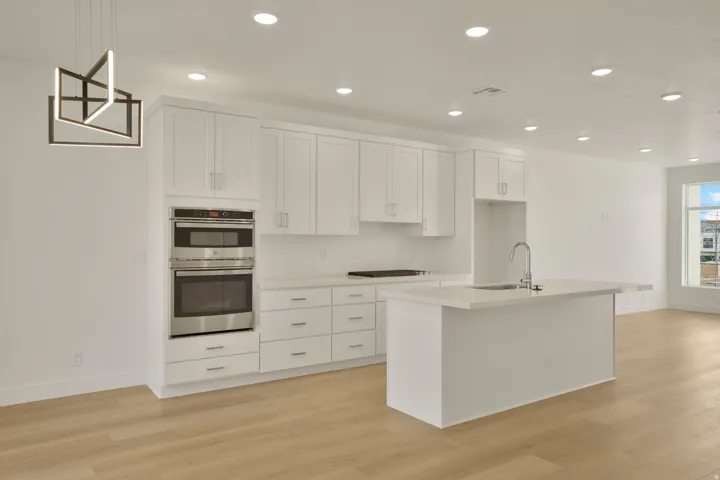 Modern kitchen featuring white shaker-style cabinetry, a built-in stainless steel oven and microwave, light-toned wood-finish flooring, a contemporary island with an integrated sink, and recessed ceiling lighting